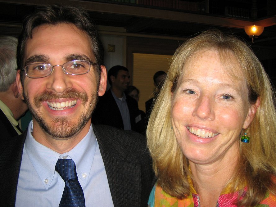 A man in a suit and tie wearing glasses and a blond woman smile at the camera