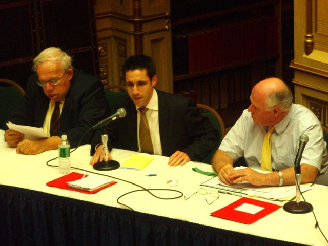 A young man in a suit and tie with glasses speaks into a microphone while sitting at a conference panel table surrounded by two other men